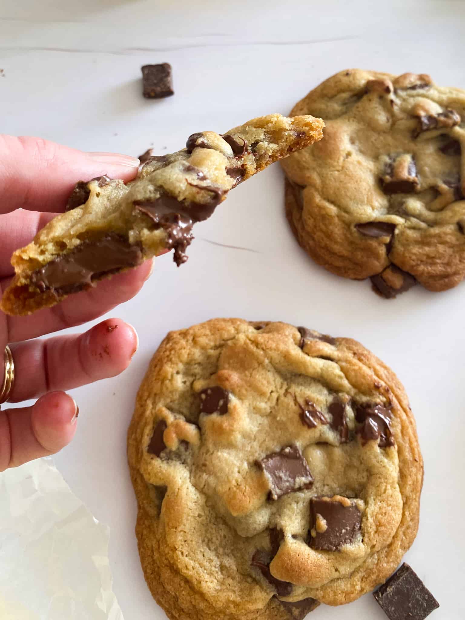 the inside of a perfectly baked gooey chocolate chip cookie (held by a hand) with whole cookies and chocolate chips in the background