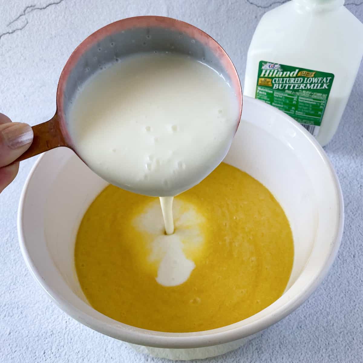 pouring buttermilk into a bowl with melted butter, eggs, sugar, and olive oil with a carton of Hiland Dairy buttermilk in the background